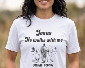May include: A white short-sleeved t-shirt featuring black text that reads Jesus He walks with me and John 10:14. Below the text is a black and white illustration of a figure holding a staff walking on a path with a sheep in a landscape.