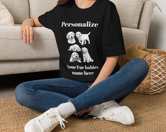 May include: A black short-sleeved t-shirt featuring white text that reads Personalize Your Fur babies name here, accompanied by a graphic illustration of five white golden retriever puppies in various poses arranged in a central cluster.