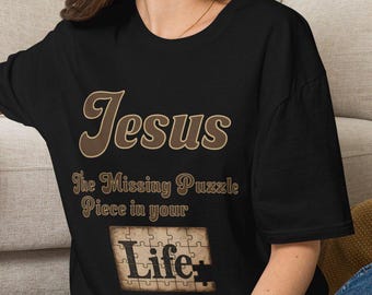 May include: A black short-sleeved t-shirt featuring the text "Jesus The Missing Puzzle Piece in your Life" in a brown font. Below the text is a graphic of a tan puzzle with the word "Life" printed in black, including one missing piece.