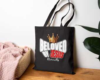 May include: A black tote bag featuring a gold crown graphic above the white text "BELOVED BY" and the red text "JESUS" with the word "Eternally" in white script underneath. The bag has two black shoulder straps and a rectangular shape.