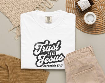 May include: A white short-sleeved t-shirt featuring black distressed-style lettering that reads Trust In Jesus and Jeremiah 17:7. The shirt has a crew neckline and a Comfort Colors brand label visible at the inner collar area.