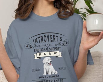 May include: A slate blue t-shirt featuring a graphic of a white puppy. The text reads: INTROVERT'S EMOTIONAL SUPPORT FLAW HIS CUTENESS RUINED MY PLANS TO STAY INVISIBLE. The design includes decorative black scrollwork, a heart, and a banner element.