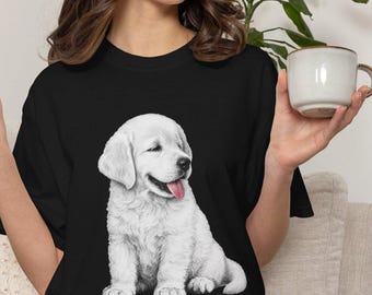 May include: A black short-sleeved t-shirt featuring a detailed graphic illustration of a white golden retriever puppy sitting down with its tongue out. The puppy is rendered in shades of white and gray, contrasting against the solid black fabric.