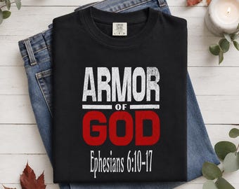 May include: A black short-sleeved t-shirt featuring the white distressed text ARMOR OF, the red distressed text GOD, and the white text Ephesians 6:10-17 printed on the front. The shirt has a crew neckline and a visible interior brand label.