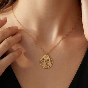 May include: A gold-colored necklace with a pendant featuring a geometric design and a small initial. The necklace is worn on a person wearing a black top. The pendant is circular with a detailed pattern.