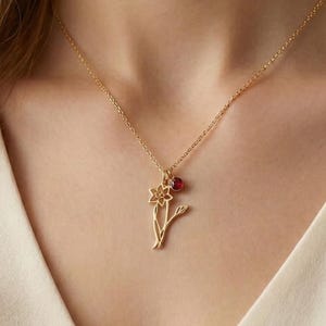 May include: A gold-colored necklace with a floral pendant. The pendant features a flower and a small red gemstone. The necklace is worn against a cream-colored top, creating a contrast with the gold chain and pendant.