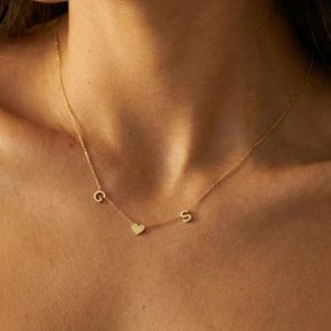 May include: A delicate gold necklace with three charms: the letter "G", a small heart, and the letter "S". The necklace is worn around the neck, resting on the skin. The overall aesthetic is minimalist and elegant, suitable for everyday wear.