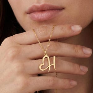 May include: Gold-colored necklace with a pendant featuring the letter "H" intertwined with a heart shape. The pendant hangs from a delicate chain. The necklace is displayed against a soft, neutral background.