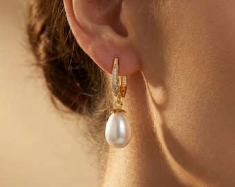 May include: A gold hoop earring featuring a small row of clear stones along the front edge and a smooth, white, teardrop-shaped pearl pendant hanging from the base of the hoop. The earring is secured through the earlobe with a hinged clasp.