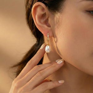 May include: A gold-colored earring with a small row of clear stones at the top and a dangling white pearl. The earring is a hoop style with a lever back closure. The pearl is teardrop-shaped.