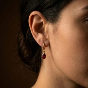 May include: Gold hoop earring with a teardrop-shaped, deep red gemstone. The earring is worn on the ear of a person with dark hair and skin. The earring is a simple, elegant design.