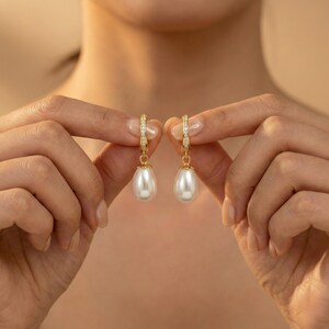 May include: A pair of gold-colored hoop earrings with a teardrop-shaped white pearl pendant. The hoops are adorned with small, clear gemstones. The earrings are held up by a person's hands against a neutral background.