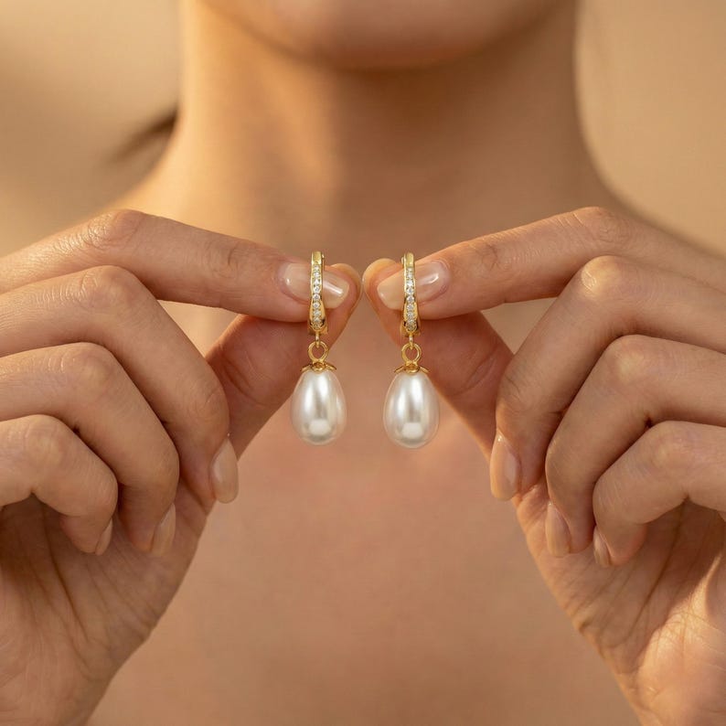 May include: A pair of gold-colored hoop earrings with a teardrop-shaped white pearl pendant. The hoops are adorned with small, clear gemstones. The earrings are held up by a person's hands against a neutral background.