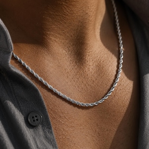 Puede incluir: Un collar de cadena de cuerda plateada. El collar se lleva alrededor del cuello y está hecho con un diseño retorcido en forma de cuerda. La persona lleva una camisa gris con botones.