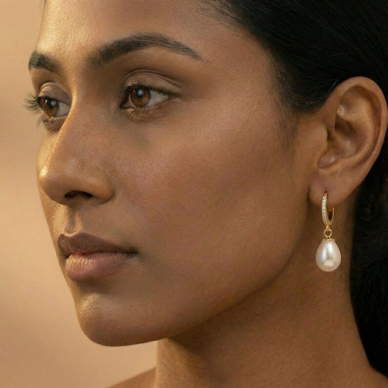 May include: A close-up of a person wearing a gold hoop earring with a white teardrop pearl pendant. The earring is encrusted with small, clear gemstones. The background is a solid, warm beige color.