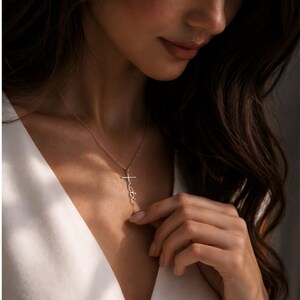 May include: A delicate gold-colored necklace featuring a cross pendant with the word "faith" written vertically. The necklace is worn against a white top, with the focus on the jewelry.