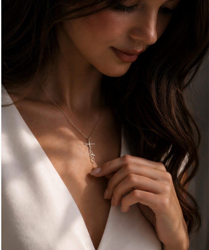 May include: A delicate gold-colored necklace featuring a cross pendant with the word "faith" written vertically. The necklace is worn against a white top, with the focus on the jewelry.
