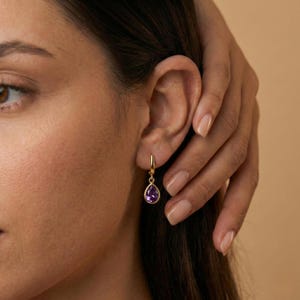 May include: Gold-colored hoop earrings with a teardrop-shaped, deep purple gemstone. The earrings are shown on a person's ear, with the gemstone hanging below the earlobe. The background is a solid, neutral color.