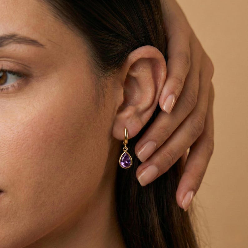 May include: Gold-colored hoop earrings with a teardrop-shaped, deep purple gemstone. The earrings are shown on a person's ear, with the gemstone hanging below the earlobe. The background is a solid, neutral color.
