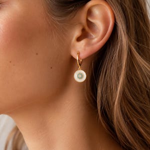 Pode incluir: Brincos de argola dourados com um pendente circular branco. O pendente apresenta um design turquesa e dourado no centro, rodeado por uma borda dourada. Os brincos são mostrados na orelha.