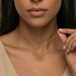 May include: A gold-colored necklace with a diamond-shaped pendant. The pendant has a textured design and hangs from a delicate chain. The necklace is worn around the neck.