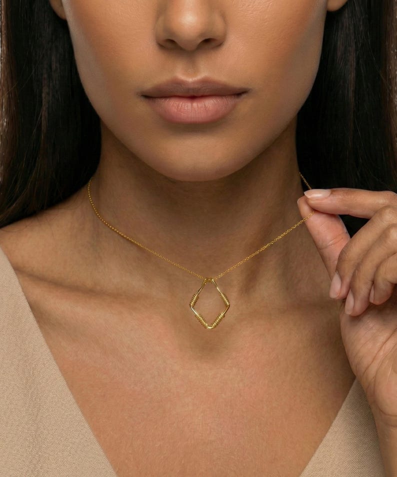 May include: A gold-colored necklace with a diamond-shaped pendant. The pendant has a textured design and hangs from a delicate chain. The necklace is worn around the neck.