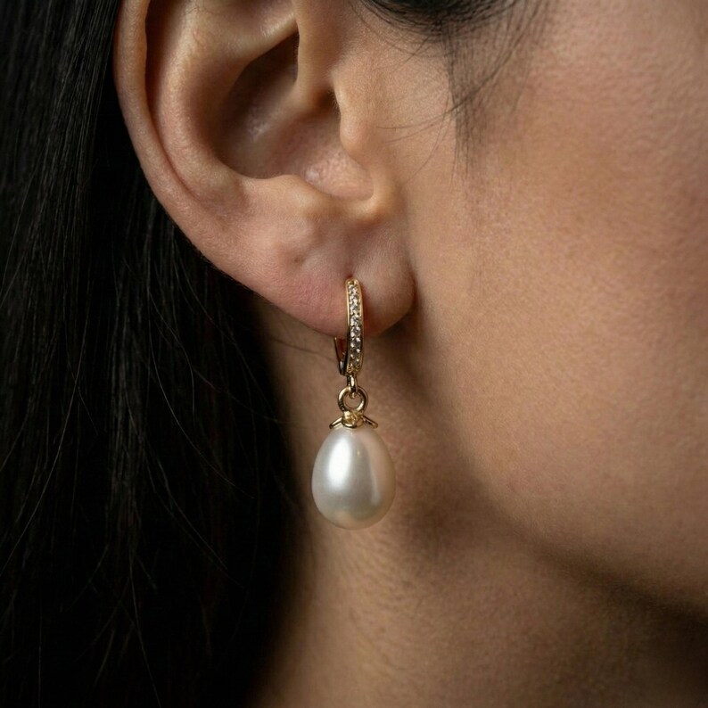 May include: A gold hoop earring with small clear stones and a dangling white teardrop-shaped pearl. The earring is attached to an ear. The background is dark.
