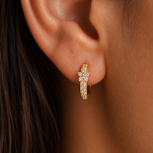Op de afbeelding: Goudkleurige oorring met een bloemmotief. De ring is bezet met kleine, heldere edelstenen. De bloem aan de bovenkant van de ring is gemaakt van heldere edelstenen. De oorring wordt gedragen op het oor van een persoon.