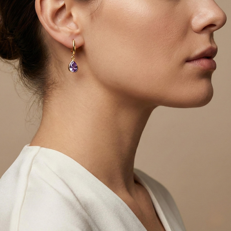 May include: Gold hoop earring with a teardrop-shaped purple gemstone. The earring is worn on the ear of a person wearing a white top. The background is a neutral beige color.