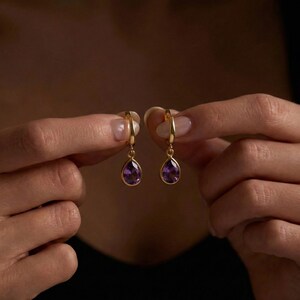 May include: A pair of gold-colored hoop earrings, each featuring a teardrop-shaped, deep purple gemstone. The earrings are held up against a dark background, showcasing their elegant design and color contrast.