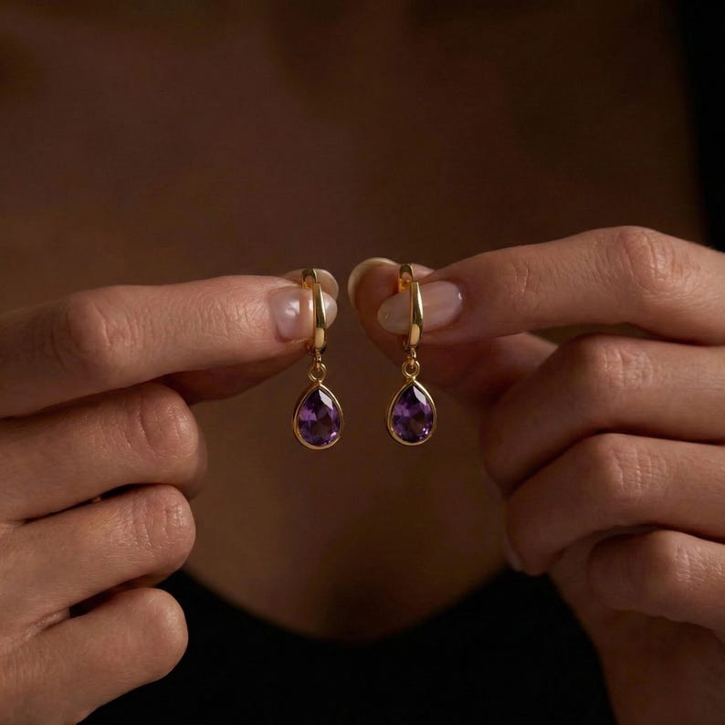 May include: A pair of gold-colored hoop earrings, each featuring a teardrop-shaped, deep purple gemstone. The earrings are held up against a dark background, showcasing their elegant design and color contrast.