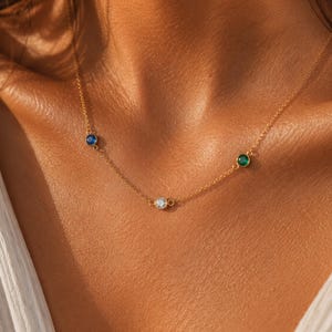 May include: A delicate gold necklace featuring three bezel-set gemstones: a blue sapphire, a clear crystal, and a green emerald. The necklace is displayed on a person's skin, highlighting the jewelry's design and color.