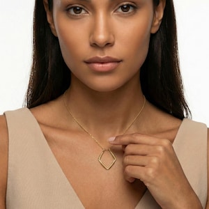 May include: A gold-colored necklace featuring a diamond-shaped pendant. The pendant is suspended from a delicate chain. The woman in the image is wearing a beige top.