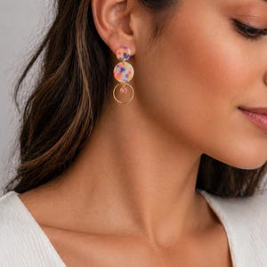 Può includere: Orecchini pendenti color oro con tre pendenti circolari colorati e maculati. I due pendenti superiori sono collegati, con un cerchio d'oro più piccolo e una gemma rosa che pende sotto. Gli orecchini sono indossati.
