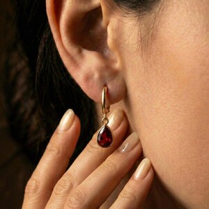 May include: A gold hoop earring with a teardrop-shaped, deep red gemstone pendant. The earring is worn on an ear, with a hand gently touching the ear. The gemstone is a rich, dark red color.