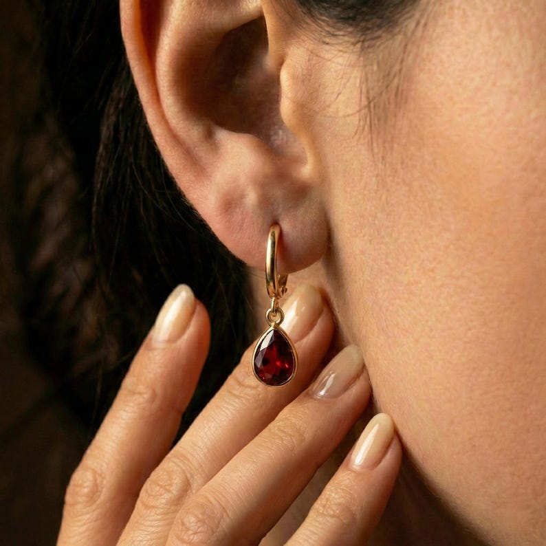 May include: A gold hoop earring with a teardrop-shaped, deep red gemstone pendant. The earring is worn on an ear, with a hand gently touching the ear. The gemstone is a rich, dark red color.