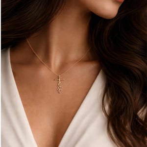 May include: A delicate gold necklace featuring a cross pendant with the word "faith" written in a cursive script. The necklace is worn against a white top, highlighting the pendant's design. The jewelry is a simple yet elegant accessory.