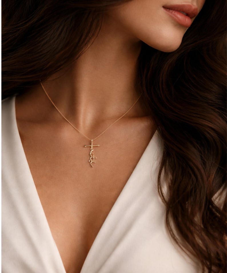 May include: A delicate gold necklace featuring a cross pendant with the word "faith" written in a cursive script. The necklace is worn against a white top, highlighting the pendant's design. The jewelry is a simple yet elegant accessory.