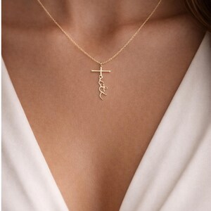 May include: A gold-colored necklace with a cross pendant. The word "faith" is written vertically on the cross. The necklace is worn against a white top.