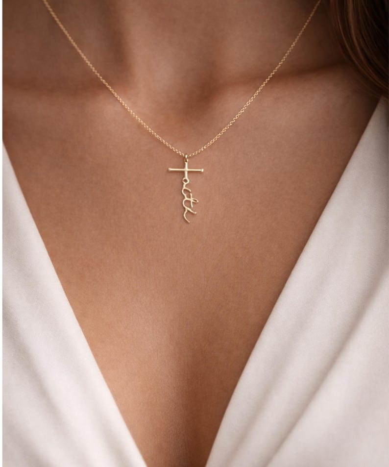 May include: A gold-colored necklace with a cross pendant. The word "faith" is written vertically on the cross. The necklace is worn against a white top.