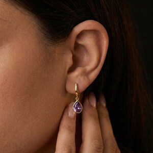 May include: A gold-colored earring with a teardrop-shaped, purple gemstone. The earring is hanging from a gold hoop. The image shows the earring on a person's ear.