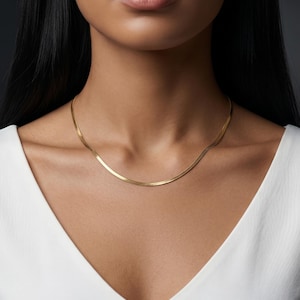 May include: A gold-colored herringbone chain necklace. The necklace is worn around the neck and rests on a white v-neck top. The chain has a flat, woven design and a subtle shine. The background is a dark gray.