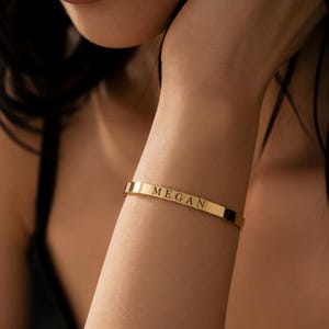 Puede incluir: Una pulsera tipo brazalete de metal dorado con un acabado liso y pulido. La pulsera presenta el nombre MEGAN grabado en una fuente serif clara en mayúsculas, centrado en la superficie frontal de la banda. La pieza tiene un perfil fino.