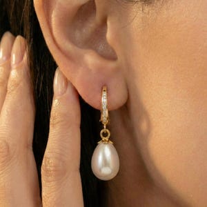May include: A gold hoop earring with small clear stones and a teardrop-shaped white pearl. The earring is hanging from an ear. The gold has a shiny finish.