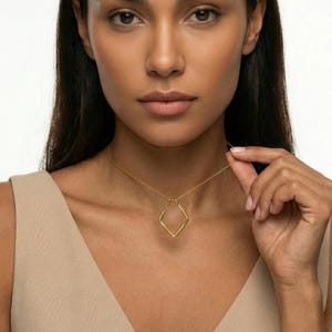 May include: A gold-colored necklace featuring a diamond-shaped pendant. The pendant has a textured surface and hangs from a delicate chain. The model is wearing a beige top.