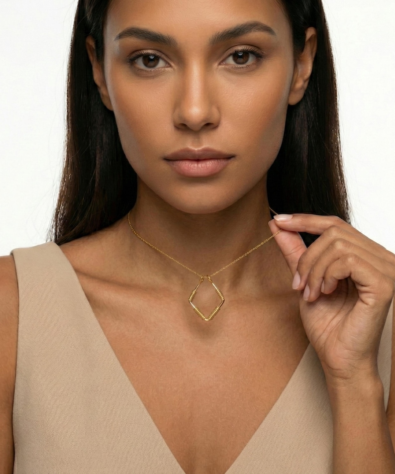 May include: A gold-colored necklace featuring a diamond-shaped pendant. The pendant has a textured surface and hangs from a delicate chain. The model is wearing a beige top.