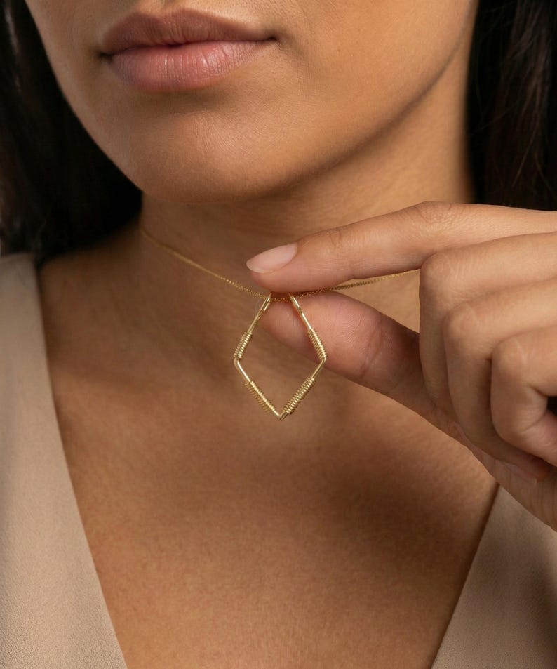 May include: A delicate gold necklace featuring a geometric diamond-shaped pendant. The pendant is crafted with thin gold wire, with wrapped detailing at the corners. The necklace is shown against a neutral background.