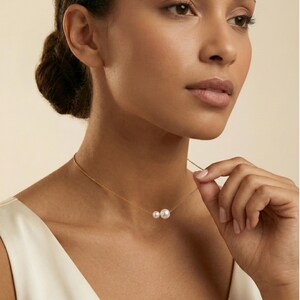 May include: A delicate gold necklace featuring two white pearls. The necklace is worn around the neck. The woman is wearing a white dress. The background is a soft, neutral color.