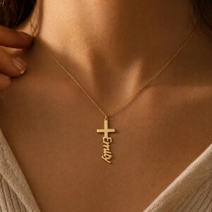 May include: A gold-colored necklace featuring a thin chain and a vertical cross pendant. The name Emily is written in a cursive script font integrated into the lower portion of the cross, extending downward from the horizontal bar of the cross.