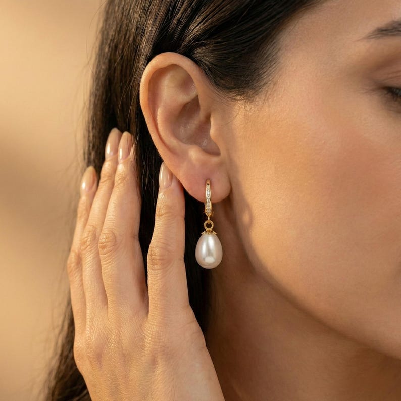 May include: A gold hoop earring with a dangling white teardrop-shaped pearl. The earring is adorned with small, clear gemstones. The image shows the earring on a person's ear.